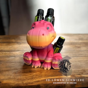 Op de afbeelding: Een roze en oranje 3D-geprint dinosaurusbeeldje met vier sleuven voor essentiële olieflessen. De dinosaurus zit op een houten oppervlak. De flessen zijn gelabeld met het woord "Terra" en de naam van de essentiële olie.