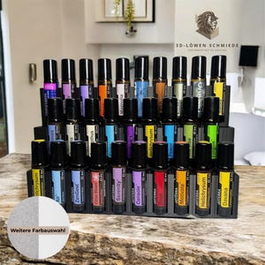 Könnte beinhalten: Ein schwarzer Holzständer mit 24 durchsichtigen Glas-Rollflaschen gefüllt mit ätherischen Ölen. Die Flaschen sind mit Namen wie "ZenGest", "Serenity", "Rose" und "Helichrysum" beschriftet. Der Ständer steht auf einer Holzoberfläche. Der Text "Weitere Farbauswahl" ist auf dem Ständer sichtbar.
