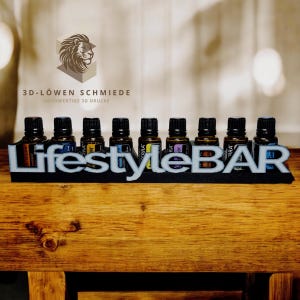 Op de afbeelding: Een zwarte 3D-geprinte standaard met de tekst "Lifestyle BAR" in witte letters. De standaard houdt tien kleine glazen flesjes met etiketten waarop "Terra" staat en met verschillende gekleurde vloeistoffen.