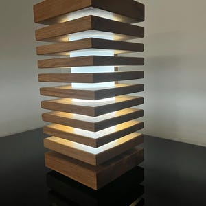May include: A rectangular table lamp constructed from stacked horizontal wooden slats with a natural brown grain. A central white cylindrical light diffuser is positioned between the wooden layers, emitting a soft glow from within the structure.