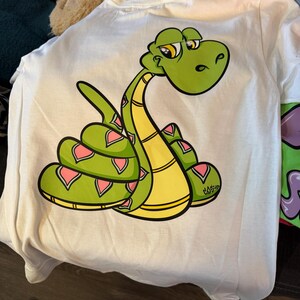 May include: White t-shirt with a cartoon snake design. The snake is green with yellow and pink details. The snake is coiled and has a relaxed expression. The shirt is made of a soft, comfortable material.