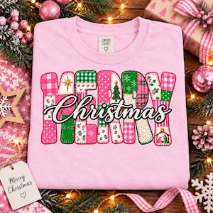 May include: A light pink crewneck sweatshirt featuring the text Merry Christmas in a patchwork-style font. The letters are filled with various patterns including pink and green plaid, polka dots, floral prints, and a small nativity scene graphic.