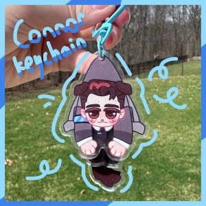 May include: A clear acrylic keychain featuring a cartoon illustration of a character with dark hair and a suit. The keychain has the text "Connor keychain" in blue cursive. The background is a blurred green lawn.
