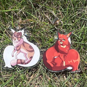 Op de afbeelding: Twee heldere acryl sleutelhangers met cartoon kat illustraties. Eén sleutelhanger heeft een bruin en bruin kattenontwerp, de andere een rood kattenontwerp. Beide sleutelhangers hebben goudkleurige sluitingen en liggen op groen gras.