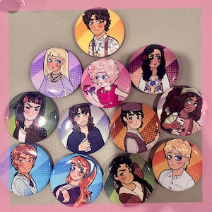 May include: A collection of round, colorful buttons featuring various cartoon character illustrations. Each button has a unique design with different characters, backgrounds, and color schemes. The buttons are arranged on a light gray surface with a pink border.