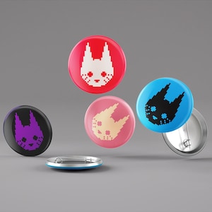 Könnte beinhalten: Vier runde Pins mit verschiedenen Hintergrundfarben und einem pixeligen Tiergesichtsdesign. Die Pins sind rot, blau, rosa und lila. Das Tiergesicht ist weiß auf dem roten Pin, schwarz auf dem blauen Pin, cremefarben auf dem rosa Pin und lila auf dem lila Pin.