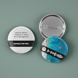 Könnte beinhalten: Zwei runde Knöpfe mit weißem Hintergrund und schwarzem Text. Der obere Knopf zeigt "Spotify Album, Playlist oder Song Art im Hintergrund" und "Spotify Code hier". Der untere Knopf zeigt eine blaue und weiße Strandkulisse mit einem rosa Flamingo und einem Spotify-Code.