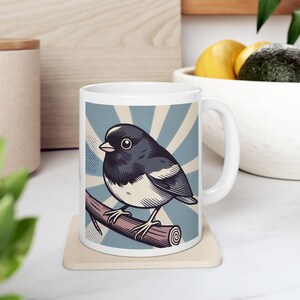May include: White ceramic mug with a black and white illustration of a bird perched on a branch. The background is a blue and white radial pattern.