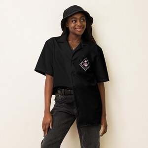 May include: A black button-up shirt with short sleeves and a patch on the left chest. The patch is a pink and white diamond with the text "Ghost Summer" inside. The shirt is worn by a person wearing a black bucket hat and black jeans.
