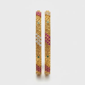 Può includere: Due braccialetti d'oro con intricati motivi floreali e pietre preziose incastonate. I braccialetti presentano sezioni alternate di pietre rosse, bianche e verdi, creando un design vibrante e ornato. L'oro ha una finitura opaca.
