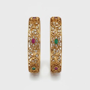Peut inclure: Deux bracelets dorés ornés de motifs floraux et de volutes complexes. Chaque bracelet est orné de petites pierres précieuses transparentes et de pierres ovales plus grandes, rouges et vertes. Les bracelets sont présentés sur fond blanc.