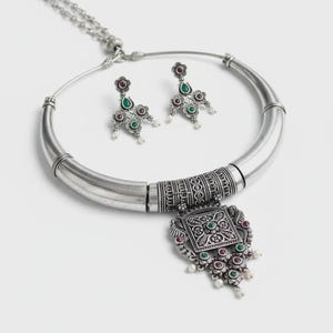 Può includere: Un set di collana e orecchini argentati. La collana ha un ampio colletto curvo con un pendente dettagliato con accenti rossi, verdi e bianchi. Gli orecchini abbinati hanno un design floreale con pietre dai colori simili.