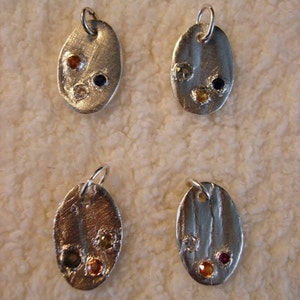 May include: Four silver oval pendants with different colored gemstones. Each pendant has three gemstones set in the metal. The gemstones are red, blue, yellow, and orange.