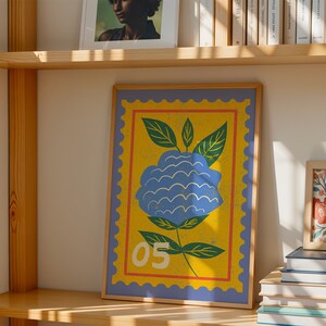 Puede incluir: Una impresi&oacute;n art&iacute;stica enmarcada con una ilustraci&oacute;n de una flor azul sobre un fondo amarillo, que se asemeja a un sello postal. Tiene un borde azul y el n&uacute;mero "05". La obra de arte se exhibe en una estanter&iacute;a de madera, junto con otros marcos y libros.