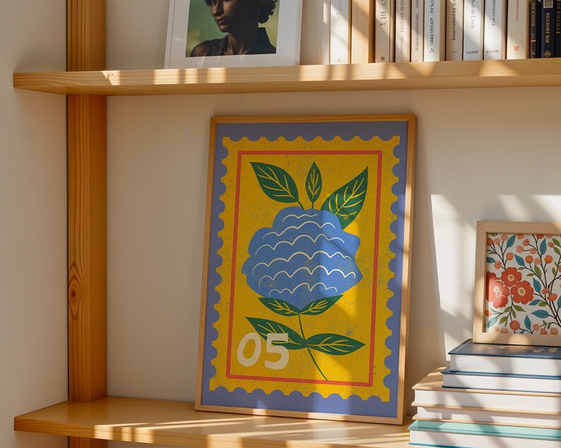 Puede incluir: Una impresi&oacute;n art&iacute;stica enmarcada con una ilustraci&oacute;n de una flor azul sobre un fondo amarillo, que se asemeja a un sello postal. Tiene un borde azul y el n&uacute;mero "05". La obra de arte se exhibe en una estanter&iacute;a de madera, junto con otros marcos y libros.
