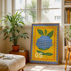 Puede incluir: Una impresi&oacute;n art&iacute;stica enmarcada con una ilustraci&oacute;n de una flor azul sobre un fondo amarillo y un borde azul. La obra de arte incluye hojas verdes y el n&uacute;mero "05". La impresi&oacute;n se exhibe en una habitaci&oacute;n con plantas y una ventana.