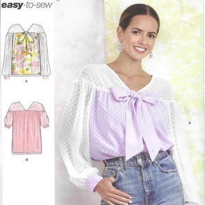 May include: A Simplicity S9921 sewing pattern envelope featuring a blouse with long sheer sleeves, a V-neckline, and a large front bow. The design includes two variations, A and B, with dotted fabric textures and a light purple color scheme.