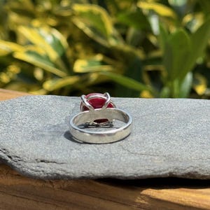 K&ouml;nnte beinhalten: Ein silberner Ring mit einem gro&szlig;en, oval geformten, roten Edelstein. Der Ring liegt auf einer grauen Steinoberfl&auml;che, mit einem verschwommenen gr&uuml;nen und gelben Hintergrund aus Bl&auml;ttern. Das Band des Rings ist glatt und poliert.