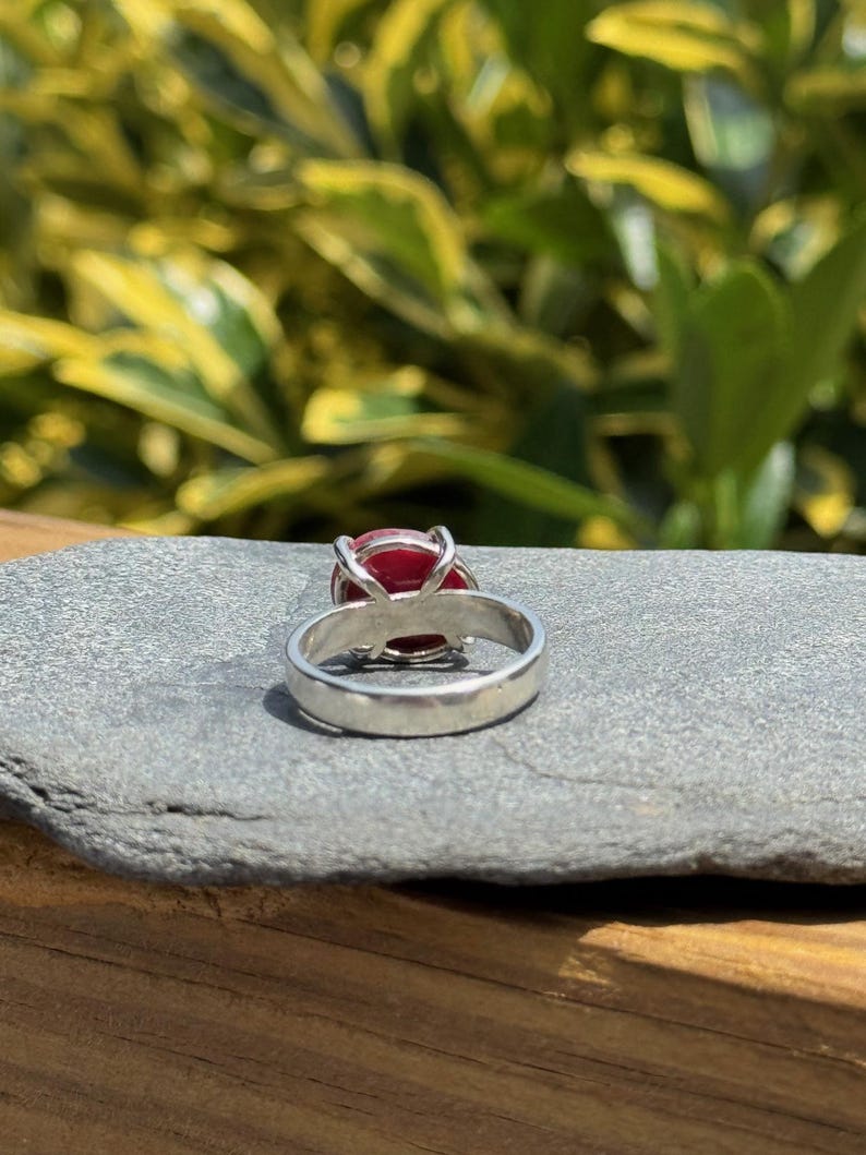 K&ouml;nnte beinhalten: Ein silberner Ring mit einem gro&szlig;en, oval geformten, roten Edelstein. Der Ring liegt auf einer grauen Steinoberfl&auml;che, mit einem verschwommenen gr&uuml;nen und gelben Hintergrund aus Bl&auml;ttern. Das Band des Rings ist glatt und poliert.