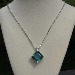 Puede incluir: Un collar de plata con un colgante cuadrado de piedra preciosa de color turquesa. El colgante está engastado en un marco de plata y cuelga de una delicada cadena. El collar se exhibe en un maniquí blanco.