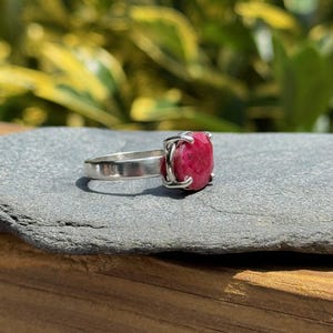 K&ouml;nnte beinhalten: Ein Silberring mit einem gro&szlig;en, quadratisch geschliffenen, tiefroten Edelstein. Der Edelstein wird von vier Krappen gehalten. Der Ring liegt auf einer grauen Steinoberfl&auml;che, mit einem verschwommenen gr&uuml;n-gelben Hintergrund.