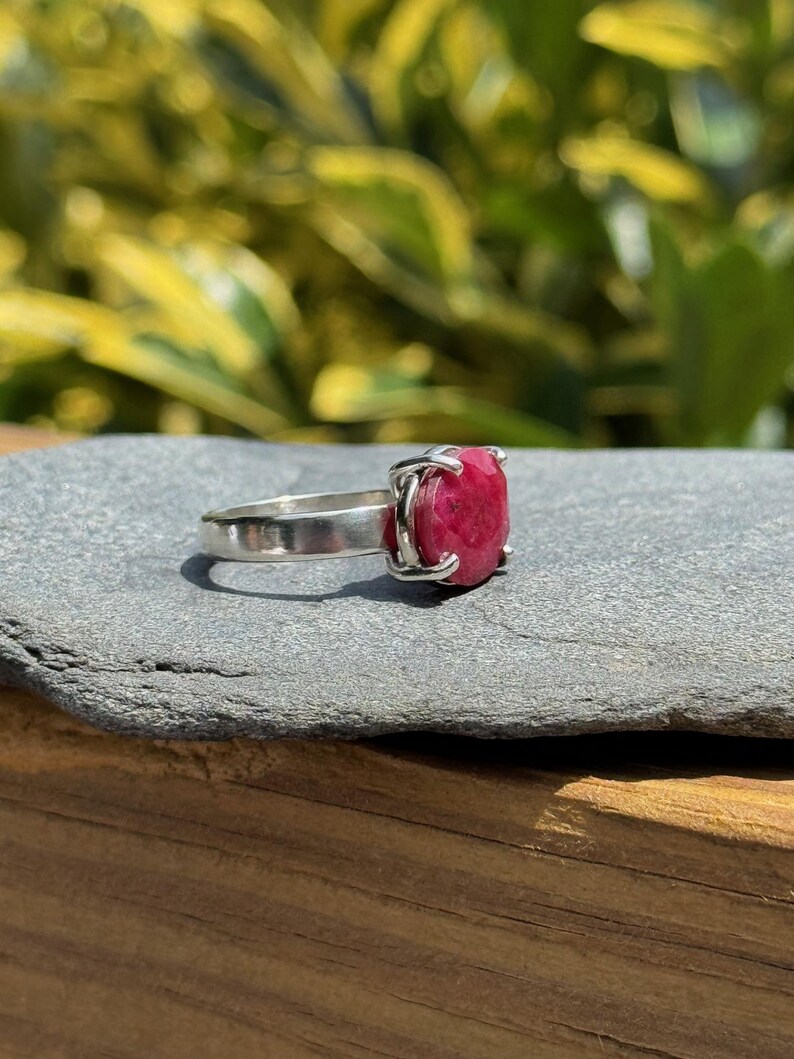 K&ouml;nnte beinhalten: Ein Silberring mit einem gro&szlig;en, quadratisch geschliffenen, tiefroten Edelstein. Der Edelstein wird von vier Krappen gehalten. Der Ring liegt auf einer grauen Steinoberfl&auml;che, mit einem verschwommenen gr&uuml;n-gelben Hintergrund.