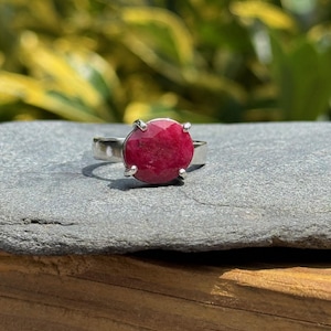 May include: A silver ring featuring a large, faceted, round ruby gemstone. The ruby is a deep, rich red color and is set in a simple, four-prong setting. The ring is displayed on a gray stone surface with a blurred green background.