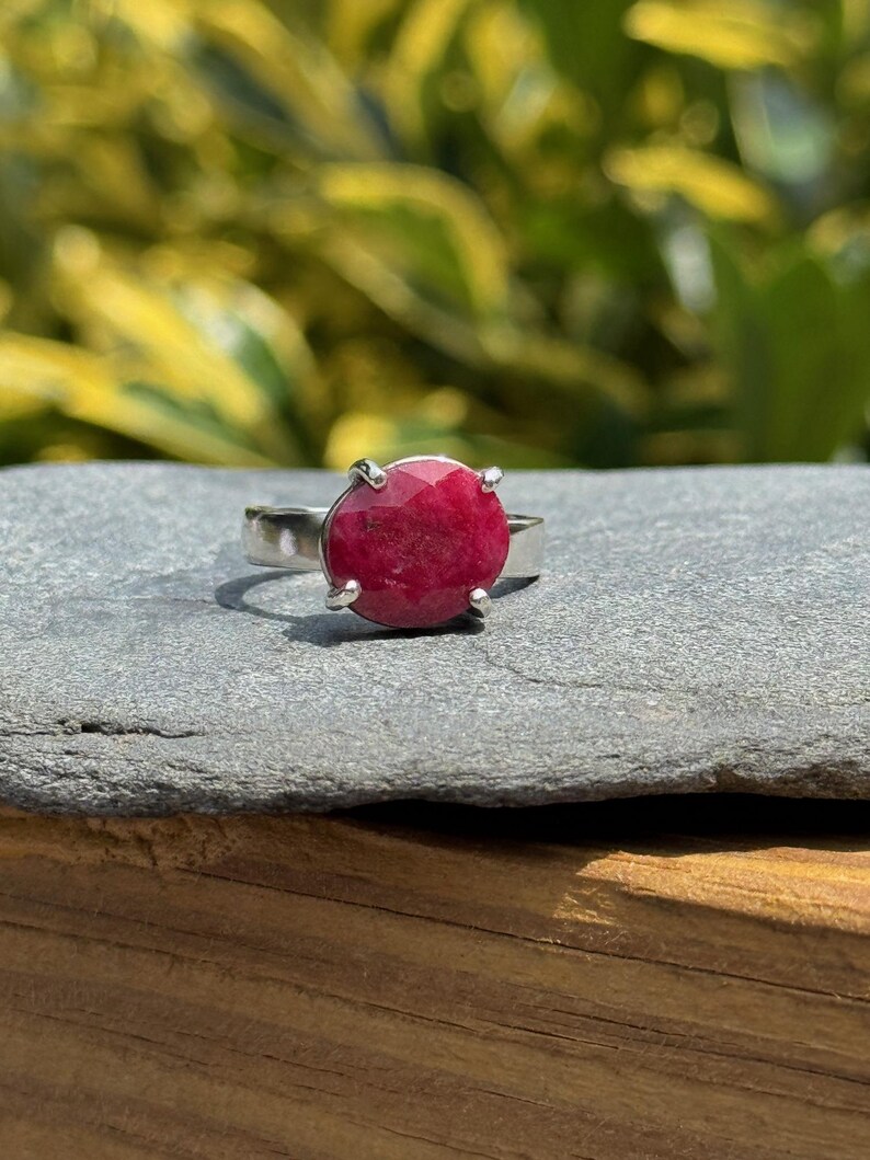 K&ouml;nnte beinhalten: Ein Silberring mit einem gro&szlig;en, runden, facettierten Rubin-Edelstein. Der Rubin hat eine tiefe, satte rote Farbe und ist in einer einfachen Vier-Zacken-Fassung gefasst. Der Ring wird auf einer grauen Steinoberfl&auml;che mit verschwommenem gr&uuml;nem Hintergrund gezeigt.