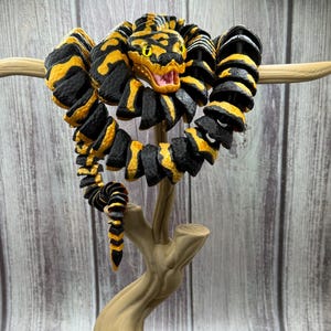 May include: A segmented, articulated snake figurine featuring a black and yellow patterned body with a detailed head, open mouth, and yellow eyes, coiled around a tan, tree-stump-shaped display stand with a textured, root-like base.