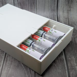 May include: A white rectangular box with a hinged lid, partially open to reveal several cylindrical items. The items are white with red and green accents, and the number "15" is visible on each. The box is on a wooden surface.