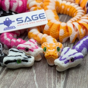 May include: A collection of articulated 3D-printed snake toys in various colors including orange, purple, pink, and black with white accents. Each snake features detailed scales and inset eyes. A white rectangular sign reads SAGE PRINTS AND DESIGN.