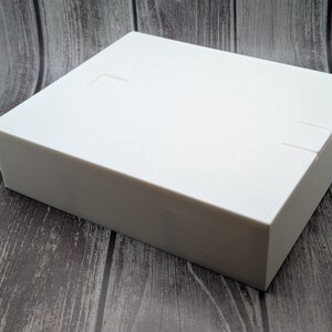 May include: A white rectangular box with a lid, set against a wooden background. The box has a smooth surface and appears to be made of a solid material. There are small indentations on the lid and side of the box.