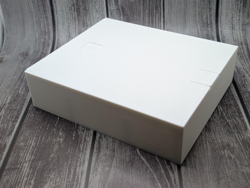 May include: A white rectangular box with a lid, set against a wooden background. The box has a smooth surface and appears to be made of a solid material. There are small indentations on the lid and side of the box.