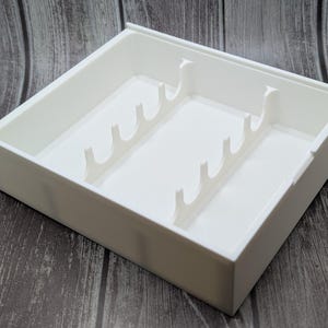 May include: A white plastic organizer tray with multiple dividers. The tray is rectangular with a raised edge and is designed to hold various items. The dividers are shaped like curved hooks, creating separate compartments within the tray.