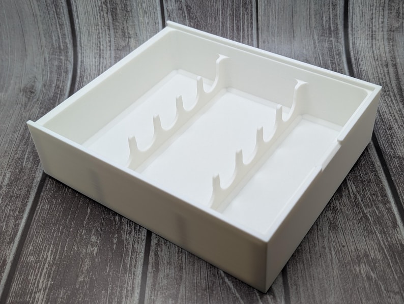 May include: A white plastic organizer tray with multiple dividers. The tray is rectangular with a raised edge and is designed to hold various items. The dividers are shaped like curved hooks, creating separate compartments within the tray.