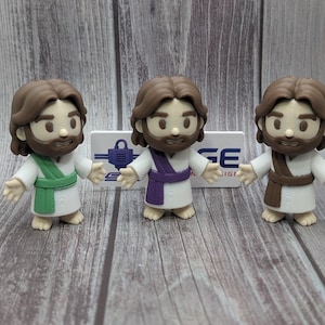 May include: Three small figurines of a person with brown hair and a beard, each wearing a white robe with a different colored sash in green, purple, or brown. The figures have outstretched arms and are positioned in front of a white sign with blue text.