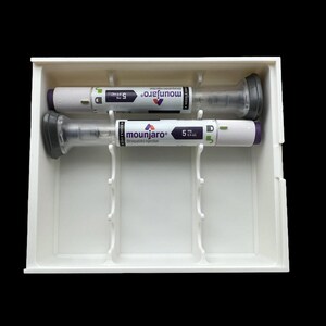 May include: Two Mounjaro prefilled injection pens, 5 mg dose, are displayed in a white plastic tray. The pens are white with gray and purple accents. The tray has dividers to hold the pens securely. The background is black.