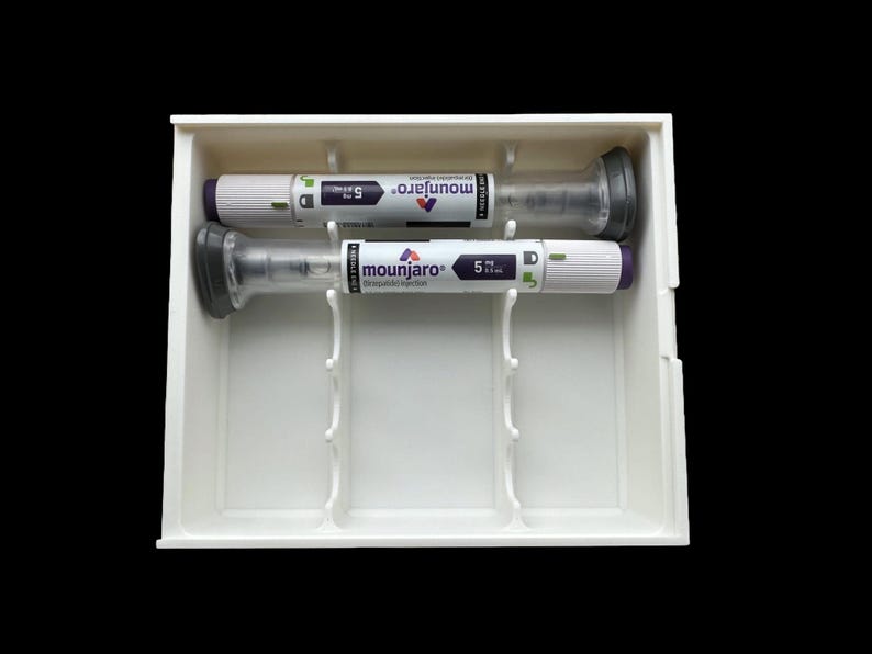 May include: Two Mounjaro prefilled injection pens, 5 mg dose, are displayed in a white plastic tray. The pens are white with gray and purple accents. The tray has dividers to hold the pens securely. The background is black.