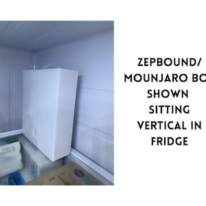 May include: A white rectangular box labeled ZEPBOUND/MOUNJARO is shown vertically inside a refrigerator. The box is the primary focus, with the fridge interior visible in the background. The box is sitting on top of other items.