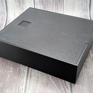 May include: A black, square box with a textured lid and a recessed square detail. The box has a matte finish and a simple, modern design. The box is sitting on a wooden surface.