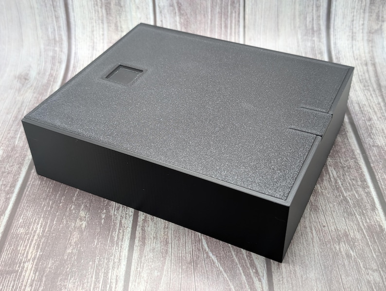 May include: A black, square box with a textured lid and a recessed square detail. The box has a matte finish and a simple, modern design. The box is sitting on a wooden surface.