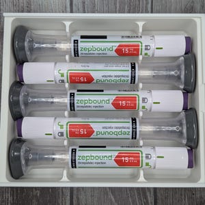May include: A white tray holds five pre-filled syringes. Each syringe has a clear barrel, a gray plunger, and a white body with green and red labels. The labels read "zepbound" and "15".