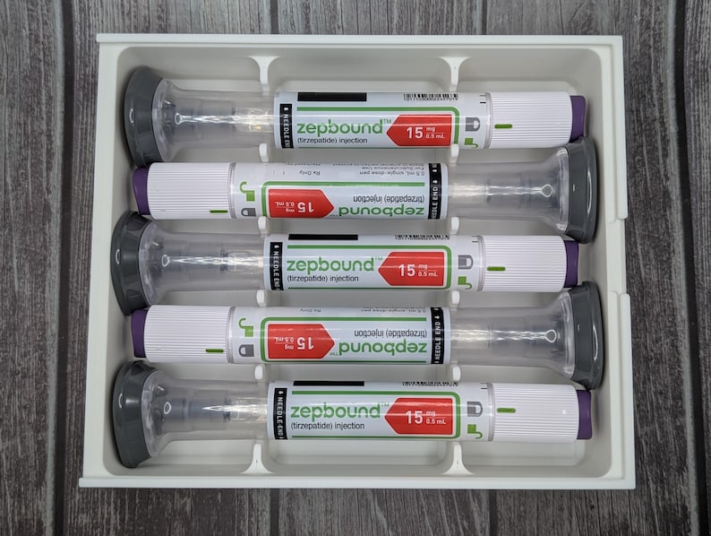 May include: A white tray holds five pre-filled syringes. Each syringe has a clear barrel, a gray plunger, and a white body with green and red labels. The labels read "zepbound" and "15".