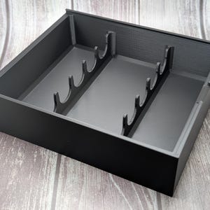 May include: A black, rectangular organizer with interior dividers. The organizer has a series of curved slots along the center, likely for holding items upright. The box is made of a matte material and sits on a wooden surface.