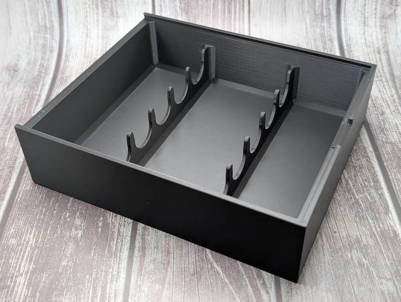 May include: A black, rectangular organizer with interior dividers. The organizer has a series of curved slots along the center, likely for holding items upright. The box is made of a matte material and sits on a wooden surface.