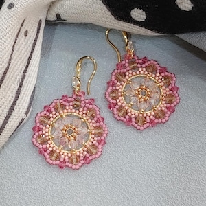 Puede incluir: Un par de pendientes hechos a mano con un diseño floral. Los pendientes tienen forma circular con un borde de cuentas rosas, un diseño central de cuentas claras y doradas, y ganchos dorados. Se muestran sobre una superficie gris claro.