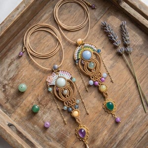 May include: Two macrame necklaces featuring tan cord, intricate knotting, and various gemstones including light blue, green, yellow, and purple cabochons. Each pendant includes small metal chains, beads, and a central stone setting.