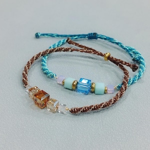 Könnte beinhalten: Zwei handgefertigte Armbänder. Eines ist braun mit transparenten und bernsteinfarbenen Würfelperlen. Das andere ist türkis mit blauen, goldenen und hellblauen Perlen. Beide Armbänder haben verstellbare Verschlüsse.
