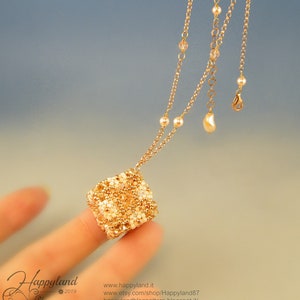 Może przedstawiać: Złoty łańcuszek z małym, koralikowym, sześciennym wisiorkiem. Wisiorek wykonany jest z złotych i białych koralików. Łańcuszek ma zapięcie karabińczykiem i mały, perłowy zawieszkę.