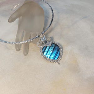 May include: A silver necklace with a heart-shaped pendant. The pendant has a blue and teal stone with a striped pattern, framed by a silver beaded border. The necklace is displayed on a clear hand.